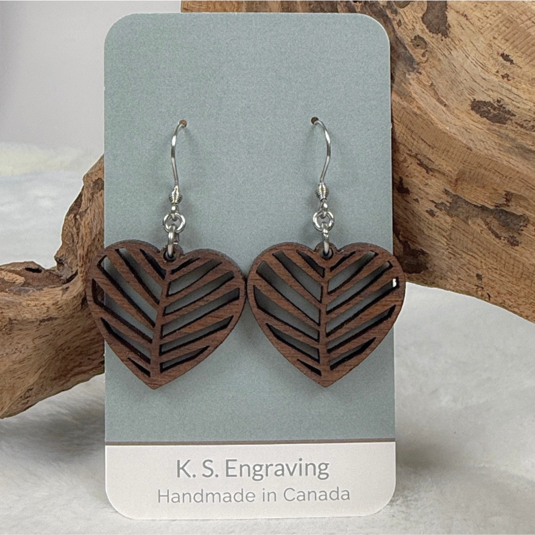 A pair of heart-shaped wooden drop earrings with a leaf cutout pattern, hanging from silver fishhook wires on a display card. The card is branded 'K.S. Engraving.