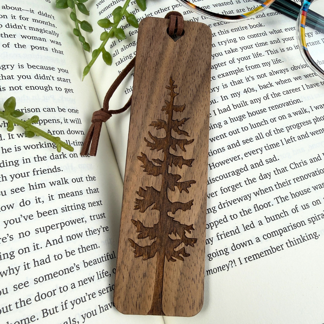 Wooden bookmark with engraved tree design on a book page, walnut wood and brown faux suede rope.