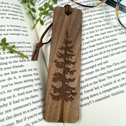 Wooden bookmark with engraved tree design on a book page, walnut wood and brown faux suede rope.