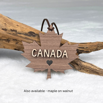 Maple and walnut Canada ornament displayed as a Canadian keepsake displayed against a soft white background with driftwood. Text at the bottom reads 'Also available - maple on walnut'.