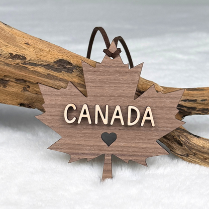 A wooden Canada maple leaf ornament with the word CANADA and a small heart cutout, hanging from brown faux suede rope and resting on a piece of driftwood.