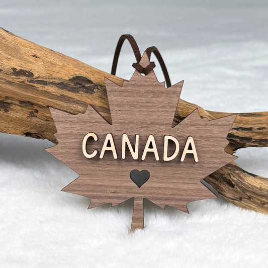 A wooden Canada maple leaf ornament with the word CANADA and a small heart cutout, hanging from brown faux suede rope and resting on a piece of driftwood.