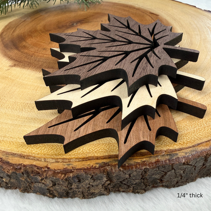 Three laser-cut wooden maple leaf coasters in different wood tones, stacked on a rustic wood slice background.