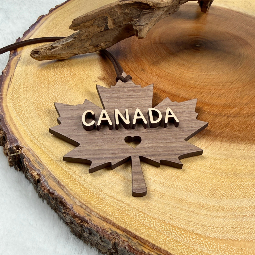 A wooden Canada maple leaf ornament with raised 'CANADA' lettering and a small heart cutout, displayed on a round wood slice background.