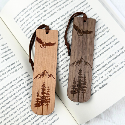 Two wooden bookmarks with laser-engraved designs of an eagle, mountains, and pine trees. Made from walnut or cherry wood with a satin finish. Includes a brown faux suede rope at the top and is displayed on an open book.