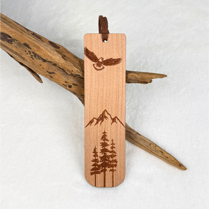 A wooden bookmark with laser-engraved designs of an eagle, mountains, and pine trees. Made from cherry wood with a satin finish. Includes a brown faux suede rope at the top and is displayed against a white textured background with driftwood.