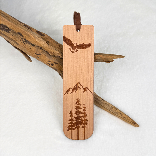 A wooden bookmark with laser-engraved designs of an eagle, mountains, and pine trees. Made from cherry wood with a satin finish. Includes a brown faux suede rope at the top and is displayed against a white textured background with driftwood.
