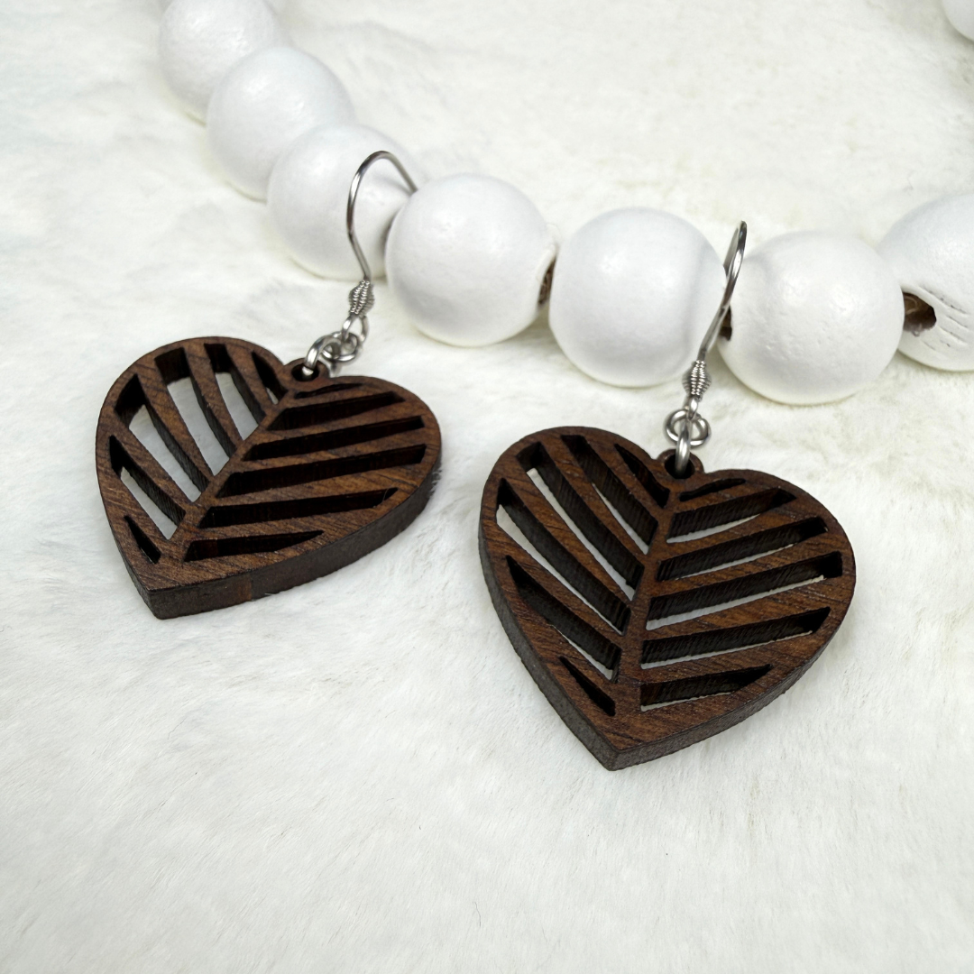 A pair of heart-shaped wooden drop earrings with a cutout pattern, hanging from silver fishhook wires. They are displayed on a white faux fur surface next to a white beads.