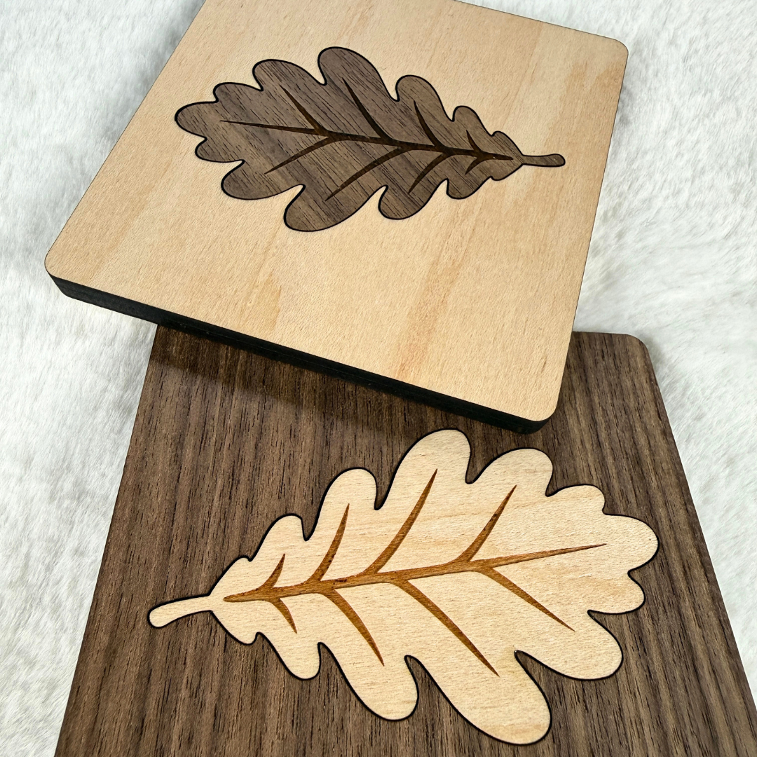 Two handmade square wood coasters featuring oak leaf inlays, one maple with walnut and one walnut with maple.