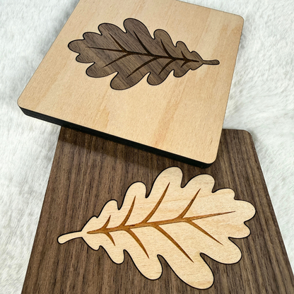 Two handmade square wood coasters featuring oak leaf inlays, one maple with walnut and one walnut with maple.