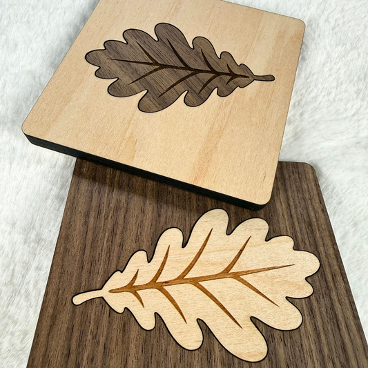 Two handmade square wood coasters featuring oak leaf inlays, one maple with walnut and one walnut with maple.