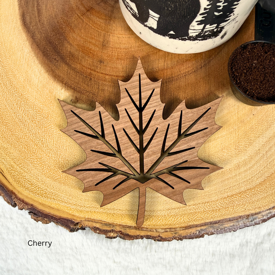 A cherry wood cutout leaf coaster with laser-cut leaf veins, displayed on a rustic wood slice coaster with a ceramic mug and coffee beans in the background.