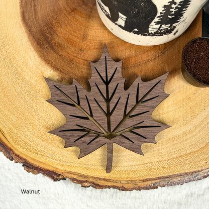 A walnut wood cutout in the shape of a maple leaf with detailed vein patterns, displayed on a rustic wood slice coaster. 