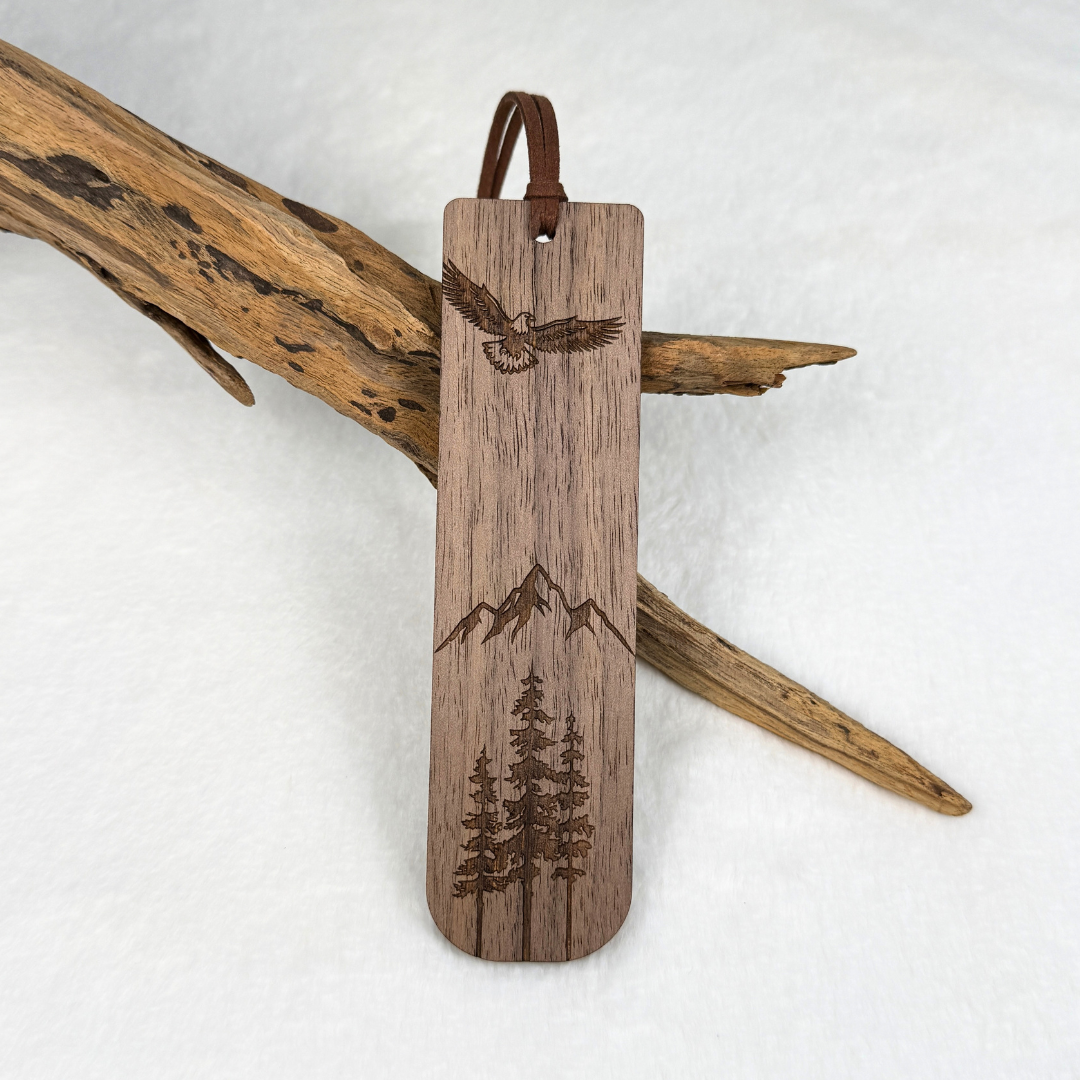 A wooden bookmark with laser-engraved designs of an eagle, mountains, and pine trees. Made from walnut wood with a satin finish. Includes a brown faux suede rope at the top and is displayed against a white textured background with driftwood.