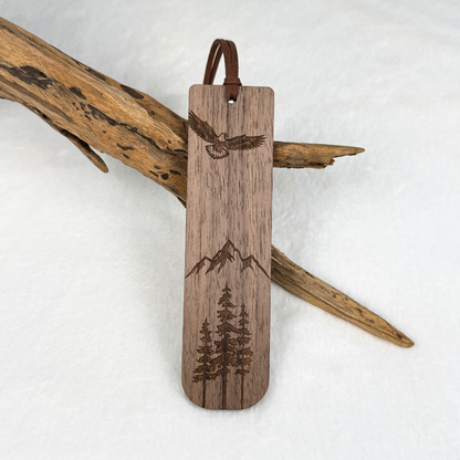 A wooden bookmark with laser-engraved designs of an eagle, mountains, and pine trees. Made from walnut wood with a satin finish. Includes a brown faux suede rope at the top and is displayed against a white textured background with driftwood.