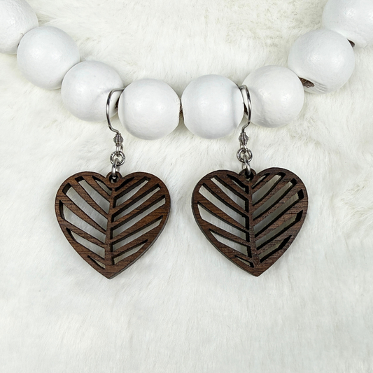 A pair of dangling heart-shaped wooden earrings with cutout leaf patterns, hanging from a white beaded necklace on a white faux fur background.