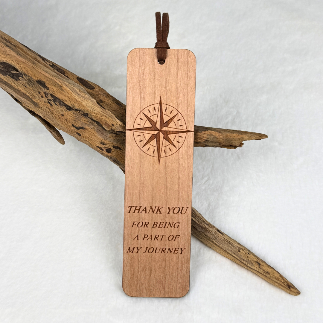 A cherry wooden bookmark with rounded corners, featuring an engraved compass  and the text 'THANK YOU FOR BEING A PART OF MY JOURNEY'. The bookmark includes a brown faux suede rope and is displayed against a white background with decorative driftwood.