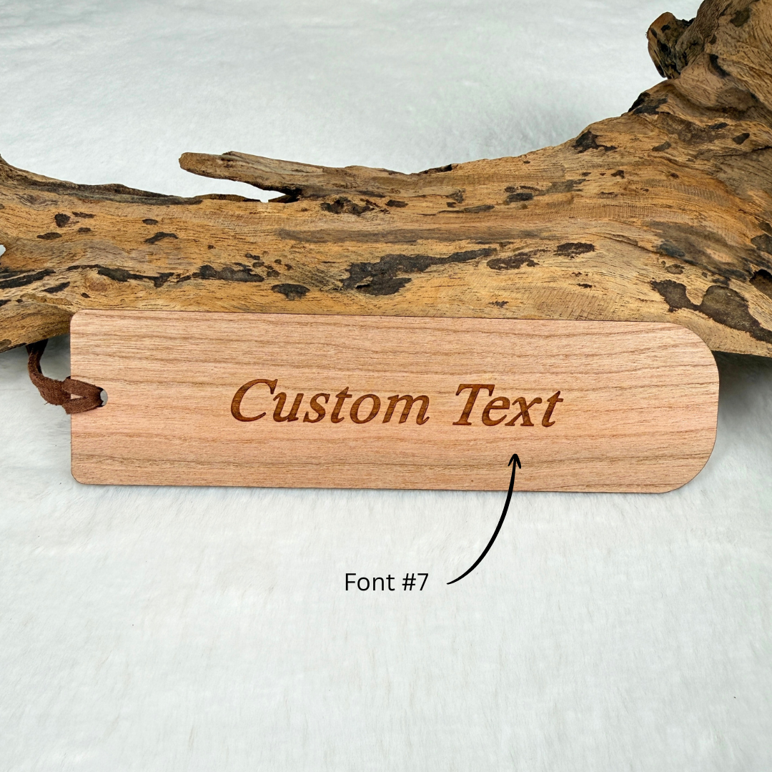 A wooden bookmark with optional personalized custom text on the back. Made from walnut or cherry wood with a satin finish. Includes a brown faux suede rope at the top and is displayed against a white textured background with driftwood.