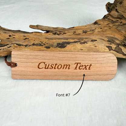 A wooden bookmark with optional personalized custom text on the back. Made from walnut or cherry wood with a satin finish. Includes a brown faux suede rope at the top and is displayed against a white textured background with driftwood.
