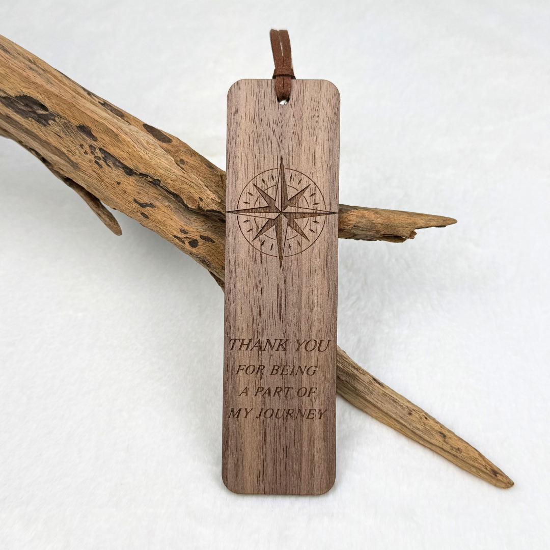 A walnut wooden bookmark with rounded corners, featuring an engraved compass  and the text 'THANK YOU FOR BEING A PART OF MY JOURNEY'. The bookmark includes a brown faux suede rope and is displayed against a white background with decorative driftwood.