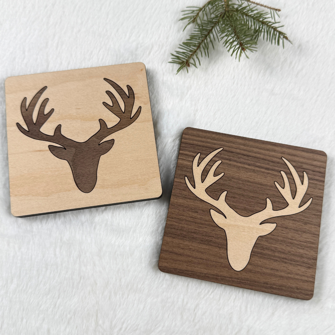 Maple and walnut wood coasters with contrasting deer head and antler inlays beside an evergreen sprig.