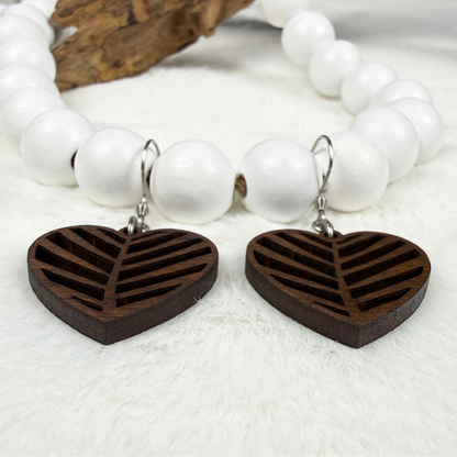 A pair of dark brown wooden heart-shaped drop earrings with laser-cut patterns, hanging from stainless steel hooks from white beads on a white textured surface.